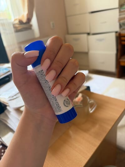 A person's hand with light pink, square-shaped nails holds a blue and white glue stick with text in multiple languages. The background is blurred and includes a desk with papers, a mug, and stacks of white boxes. There is a window with sheer curtains and what appears to be a stack of books in the far background. The scene is indoors.