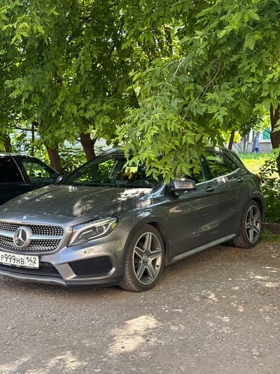 A gray Mercedes-Benz GLA-Class SUV is parked on a gravel surface under a leafy tree. The car's license plate is visible and reads "P999HB 142". The car is angled towards the viewer from the front left. In the background, a person is walking in front of a building. The scene appears to be in a residential area in Belovo, Russia, given the style of the license plate and the surroundings.