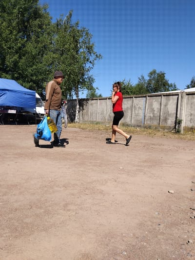 In Irshansk, Ukraine, on a clear, sunny day, a man and a woman are depicted in a sparsely populated outdoor area. The ground is covered in light brown dirt with scattered small stones and debris. In the background, a grey concrete wall runs horizontally across the scene, with trees and foliage visible beyond it.
The man, positioned on the left side of the frame, wears a brown long-sleeved shirt, blue jeans, and a dark hat. He is carrying a large bright blue plastic bag in his left hand, which contains a large yellow plastic bottle. His right hand is partially obscured by the bag. He appears to be walking.
To the right of the man, the woman is walking away from the camera. She is wearing a red short-sleeved t-shirt and black shorts. Her right hand is raised to her face, and she is looking to her left. She is wearing black slip-on shoes.
Further to the left, a blue tent is visible, along with a white vehicle and a folding table, suggesting a possible market or roadside setup. The overall scene conveys an ordinary day outdoors with two individuals in close proximity.