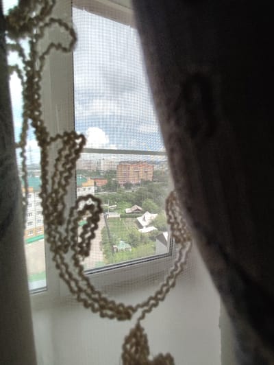 The image displays a beaded string hanging in front of a window overlooking a city. The beads are light-colored and arranged in a coiled pattern. Through the window, a cloudy sky and a cityscape are visible. The cityscape includes several buildings of varying heights and colors, some with visible windows and balconies. Green trees and foliage are scattered throughout the urban landscape, suggesting a residential area with some greenery. A thin white mesh screen is visible on the window, and to the right, a dark curtain is partially obscuring the view. The lighting suggests it is daytime. The style of the buildings and the presence of trees and a mesh screen are consistent with elements commonly found in residential areas of cities in Russia, such as Chita.
