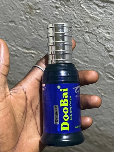 A close-up shot shows a hand holding a blue bottle of Doobai Body Spray Cologne. The bottle has "Doobai" printed in large, bright yellow letters, with "Body Spray Cologne" and "*Original*" in smaller text. A logo with a stylized bird is also visible on the bottle. The hand is dark-skinned, and a silver ring is worn on one of the fingers. The background is a textured, light-colored wall, likely indoors. The lighting suggests it is daytime. The image appears to be taken in Bauchi, Nigeria.