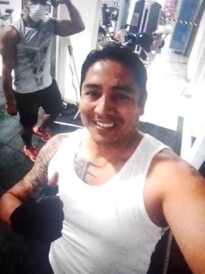 The image shows a man in a white tank top giving a thumbs up. He has a tattoo on his left arm. In the background, another man wearing a face mask and a tank top is flexing his bicep. They appear to be in a gym in the city of Santa Cruz de la Sierra, Bolivia, based on common architectural and landscaping features in the city, but could be elsewhere. Gym equipment is visible in the background. The floor appears to be dark rubber or tile.