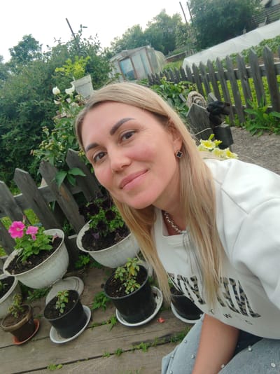 A blonde woman with fair skin and a light-colored sweatshirt is in the foreground of the image. She has on earrings and a necklace. The woman is looking at the camera and smiling, suggesting a happy or contented mood. Behind her is a wooden fence, a greenhouse, and various potted plants, indicating an outdoor setting, possibly a garden or yard. The plants are green and some have pink and dark purple flowers. The time of day appears to be daytime, given the brightness of the image, and the weather seems pleasant. The overall scene conveys a relaxed and domestic atmosphere. The woman's sweatshirt has a leopard print design with letters on it. The location is identified as Molodegniy, Russia.