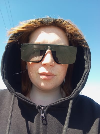 A woman with red hair and freckles is looking down at the camera while wearing a black hoodie and large sunglasses. The background is a bright blue sky, suggesting it is daytime. The photograph appears to be taken outdoors. The location of Yurgamysh, Russia is not verifiable from the image.