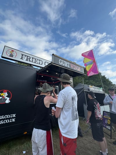 A food truck called "Fred's Fried Chicken - Gurt Wings" sits beneath a blue sky with scattered clouds. Two people wearing hats stand in front of the truck, presumably ordering. Further back, another food stall advertising "Coffee, Slushes, Shakes,..." is visible with people waiting. A colorful flag flies in the background. The side of the food truck displays its website address.