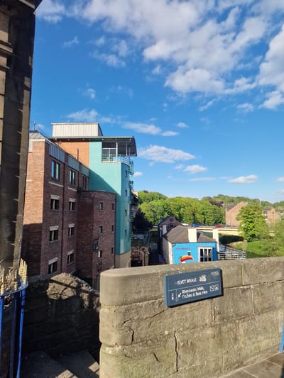 The image is a ground-level view of the Elvet Bridge in Durham, United Kingdom. The sky is blue with scattered white clouds. The bridge's stone structure occupies the right side of the image, with a sign attached to it that reads "ELVET BRIDGE," then lists options for "Riverbanks Walk, Cruises & Boat Hire." A building of varied architectural styles is visible below. It is primarily red brick with several rectangular windows, with additional sections that are teal, light gray, and copper. Adjacent to the building is a view of trees and a building that has a sign that states: "Lovely day for a..." with an umbrella image. A blue railing is visible in the lower foreground.