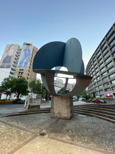 The image depicts a public art sculpture in Naucalpan de Juárez, Mexico. The sculpture is composed of blue circular shapes and rusted metal segments. The base is concrete. Behind the sculpture is a building with a large Guess billboard. Other buildings and trees are visible in the background. The sky is clear. A sidewalk, steps, and street border the sculpture. Cars are parked along the street.