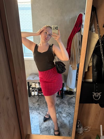 A young woman, approximately in her twenties, is captured in a mirror selfie within an indoor setting in Rossiyskiy, Russia. She holds a light-colored smartphone in her right hand, aiming it towards her reflection. Her left hand is placed on her head, and she has a slightly puckered lip expression while looking towards the phone. She wears a dark brown tank top, a short, fitted skirt with a pink and red houndstooth pattern, and black Mary Jane style shoes with two straps. A silver-toned chain necklace with a small pendant is visible around her neck, and a black shoulder bag with a chain strap is slung over her left shoulder.
The immediate surroundings visible in the reflection include a light gray textured wall, a white-framed window revealing an exterior brick wall, and a gray and white marble-patterned tiled floor. To the left of the reflection is a wooden door frame or panel. Behind the woman's legs, a small gray upholstered bench or ottoman sits above a black metal shoe rack holding several pairs of sneakers. To the right of the reflected woman, a garment rack or hooks display a red patterned jacket, a beige jacket, and a light blue shirt. A black leather-textured handbag with a distinctive "swallow" emblem, likely Pinko brand, hangs in the foreground on the right. Further to the right, on a wooden surface, a white Wi-Fi router, a small white wire-frame decorative bicycle ornament, and a white basket with papers are visible.