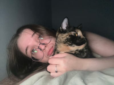 A woman with long brown hair and glasses is lying down, cradling a calico cat. The woman is wearing a silver ring on her left hand. The background is a mix of dark and light, likely indicating a room with some lighting. There is no information suggesting this image was taken in Pushchino, Russia.