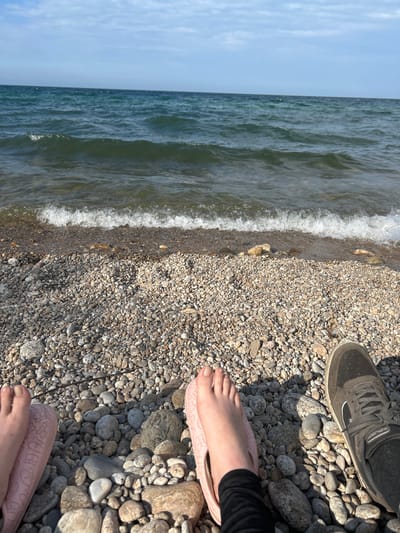 The image captures a close-up view of a rocky shoreline. In the foreground, two pairs of feet are visible. On the left, a bare foot is adorned with a light pink slipper featuring a subtle pattern. On the right, a foot is encased in a dark grey athletic shoe. The ground is covered in a dense collection of smooth, grey, and tan pebbles, with some larger stones interspersed. Gentle waves lap at the edge of the shore, creating a line of white foam. The water beyond is a deep blue-green, and the horizon is marked by a hazy sky with scattered clouds. The scene appears to be a natural setting, possibly a lake or sea, in Tolbazikha, Russia.