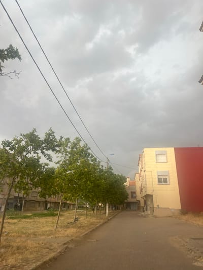 The image depicts a street scene in El Hajeb, Morocco. Two power lines stretch diagonally across the overcast sky. On the left, several trees with green foliage line a dry, grassy area. Behind the trees, an apartment building with muted tones is visible. On the right, a pale yellow building with red accents stands out, featuring white-barred windows. A paved road runs through the center of the image, leading towards the buildings. The overall atmosphere is subdued due to the cloudy weather.