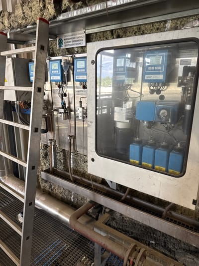 This is a forensic, matter-of-fact description of an industrial setting in Krzepielów, Poland. The scene is dominated by complex machinery and equipment, suggesting a water treatment or processing facility.
In the foreground, a metal ladder with a silver finish extends vertically along the left side of the image. It rests on a grated metal floor, which appears to be part of a walkway or platform. The floor has a diamond plate pattern and is illuminated by blue lights from below, creating a distinct visual effect.
The main subject of the image is a large, stainless steel enclosure with a glass window. Inside this enclosure, various electronic monitoring and control devices are visible. Several blue digital displays are mounted on the wall, showing readings such as "0.000" and "3.69". Some of these displays are identified with the brand "swan". Below the displays are rows of small, blue plastic bottles, each with a yellow label that also bears the "swan" logo. These bottles are connected by tubing to the machinery.
To the left of the stainless steel enclosure, a series of pipes and conduits are visible, with some electrical wiring in orange and black. There are also several vertical metal tubes with connections, suggesting a fluid or gas distribution system.
Behind the equipment, the wall is covered with a thick layer of fibrous insulation material, likely for sound or thermal purposes. This material appears somewhat disheveled.
In the background, through the glass of the enclosure and beyond the equipment, a glimpse of a natural outdoor environment can be seen, with trees and hills under a bright sky, indicating the facility is likely located in a rural or semi-rural area.
There are no people present in the image. The overall impression is one of operational industrial infrastructure, possibly related to environmental monitoring or water management, given the branding and the nature of the equipment. The presence of insulation and exposed piping suggests a functional, rather than aesthetic, design.