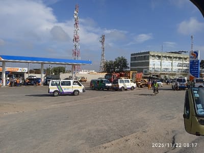 A busy street scene shows multiple vehicles including vans, cars, and trucks near a gas station under a blue awning labeled "marhaba." Several people are visible around the gas station and on the street. Two tall communication towers rise in the background beside a multi-story building. The pavement is light gray and sandy. The date "2025/11/22" and time "08:17" are stamped at the bottom of the image.