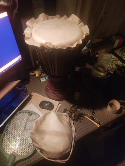 The image shows a cluttered desk or workspace in what appears to be a dimly lit room. A djembe drum with a light-colored head is prominently displayed, and next to it is a cracked replacement skin. The desk also features a computer monitor, a laptop, a mouse, a fly swatter, a water pitcher, a roll of tape, a utility knife, and various other small objects. The scene suggests a possible drum-making or repair activity. The image does not provide enough information to determine that this scene is in Ubud, Indonesia.