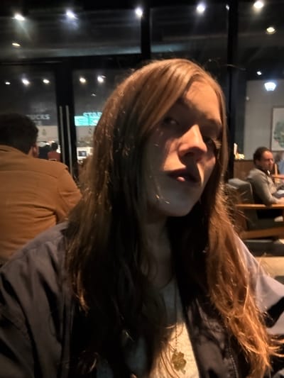 A person with long, highlighted hair is partially in focus, facing the camera. Another person sits at a table in the background, seemingly inside a Starbucks, as the Starbucks logo is visible on the storefront outside the window. A person wearing a brown shirt or jacket is also partially in view. The lighting appears dim with strong shadows on the person's face in the foreground.