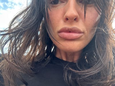 A close-up, eye-level, outdoor, daytime, selfie shot shows the lower half of a person's face and their dark, windblown hair. The person's lips are slightly parted, and their skin appears to have a light dusting of powder. A portion of a black shirt is visible at the bottom of the frame. The background consists of a bright sky with scattered clouds. The image does not contain any contextual information that would suggest a specific location within the city of Tonbridge and Malling, United Kingdom.