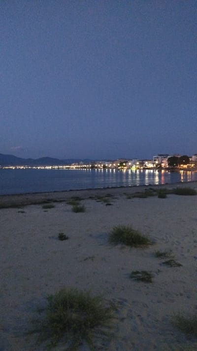 The image depicts a nighttime view of the coastline in Neoi Epivates, Greece. In the foreground, a sandy beach is visible with sparse vegetation. Beyond the beach, the calm sea reflects the lights of a town situated along the shoreline. The town appears to have low-rise buildings and is illuminated by numerous streetlights and building lights, creating a warm glow on the water. In the distance, dark hills or mountains form a backdrop against the twilight sky. The overall atmosphere is serene and peaceful, characteristic of a coastal town at dusk or night.