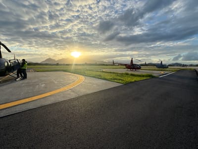 This image captures an outdoor helicopter base or helipad during either sunrise or sunset, situated within the metropolitan area of Rio de Janeiro, Brazil, against a backdrop of prominent mountains. In the immediate foreground left, two individuals stand adjacent to a dark-colored helicopter; one person, partially obscured, is near the fuselage, while the other, wearing a high-visibility yellow-green vest over dark clothing, interacts with the open cockpit door. The ground transitions from a dark asphalt surface in the very foreground to a lighter gray paved area marked with a distinct curved yellow line. Further right on the paved surface, a red Robinson R44 helicopter, clearly displaying the registration 'PR-MPT' on its tail boom, is parked. Behind it, a grey helicopter is also stationary, and multiple additional helicopters are visible in the distant background. The sky is partly cloudy with stratocumulus formations, exhibiting a mix of golden hues from the low sun and cooler gray-blue tones. The sun is a strong, bright source on the horizon, creating significant glare and silhouetting a range of large, verdant mountains. Patches of bright green grass are visible bordering the paved sections of the airfield.