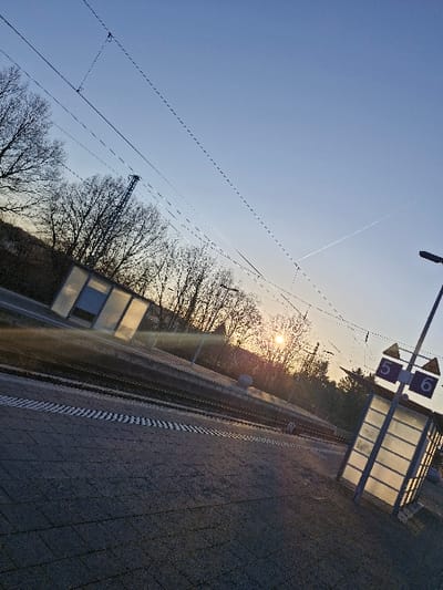 The image displays a railway platform at the Niedernhausen station in Germany, observed during either sunrise or sunset, indicated by the low sun producing a prominent lens flare and warm light hues. The platform surface is paved with dark grey rectangular blocks and features a white and black striped tactile warning strip running parallel to the tracks. Two sets of railway tracks are visible alongside the platform. Overhead catenary lines for electric trains traverse the sky. On the right side of the platform, a modern, translucent shelter made of glass and metal stands, adjacent to a signpost displaying the numbers "5" and "6", likely designating platform numbers. A black streetlight pole is also present on the right. Deciduous trees with bare branches form the background. The sky is clear, transitioning from a darker blue overhead to a lighter, warmer tone near the horizon, with a faint contrail visible in the upper right. There are no individuals, vehicles, or active operations depicted within the scene.
