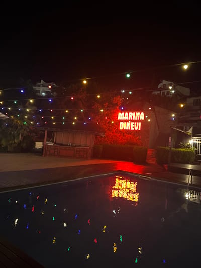 This image captures a nighttime scene at Marina Dinevi, located in Sveti Vlas, Bulgaria. The focus is on an outdoor area featuring a swimming pool and a prominent neon sign that reads "MARINA DINEVI" in red. The sign is illuminated and casts a red glow on the stone wall behind it. The swimming pool's surface reflects the colorful string lights that are strung across the scene, as well as the red neon sign, creating a vibrant display of lights. A wooden deck surrounds the pool. In the background, there are buildings and trees, also partially lit by the string lights, which are of various colors including yellow, blue, red, and green. A covered structure with patterned curtains is visible to the left of the sign. There are no people visible in the image.