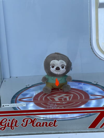 An interior view depicts a single plush toy situated within a transparent arcade prize machine. The toy is a brown, furry creature, approximately 20-25 cm in height, resembling a sloth, with a white face, dark nose, and embroidered eyes that appear partially closed with a slight smile. It is dressed in a light green striped collared shirt, khaki pants, and a prominent orange tie. The toy sits upright on a circular, reflective platform within the machine. This platform is illuminated from below, casting a bluish light, and features a central red graphic resembling a planet or orb with concentric rings and a stylized cubic shape in its core. The lower front panel of the machine is silver-toned, bearing the words "Gift Planet" in red script. A black and white warning label on the platform's edge states: "CAUTION! Do not push or lean on the glass to avoid glass breaking damage and danger." The interior walls of the machine appear light blue-grey. The scene is located within a "Gift Planet" establishment in Rishon LeZion, Israel, consistent with an arcade or entertainment venue. No human subjects are visible.