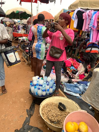 This image captures a vibrant street market scene in Nyanya, Nigeria. The focus is on several individuals interacting with a variety of goods. In the foreground, a woman with short red hair, wearing a pink t-shirt and patterned leggings, is examining something in her hands. She has a crossbody bag with a Louis Vuitton pattern. Beside her, a woman with braided hair wears a colorful, tie-dye dress and appears to be looking down.
The market is stocked with an abundance of clothing items, including stacks of t-shirts and folded garments. There are also several plastic bottles of water arranged in a tub, and a larger container filled with peanuts. In the background, more market stalls and people are visible, suggesting a bustling environment. The ground appears to be sandy or dirt, consistent with an outdoor market.
Notable details include the bright sunlight, indicating it is daytime. The overall atmosphere seems busy and active, typical of a marketplace. The image provides a glimpse into daily life and commerce in the region. There is a visible "CAUTION" label on a dark blue and white drum in the lower part of the image.