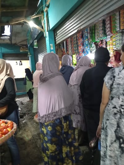 The image shows a scene from a street market in the city of Kutawaringin, Indonesia, located at coordinates -6.953 latitude, 107.524 longitude. The market is located on a street lined with shops. There are several people in the market, mostly women, all wearing traditional head coverings. They are all looking at a stall on the right side of the image. The stall has a variety of goods on display, including food and other goods. There are several bags and other objects on the ground in front of the stall. The street surface is uneven and covered in dirt. The market is busy with people moving around, buying and selling goods. The image is captured from a low angle, looking up at the people. The lighting is dim, but the scene is well lit by the shop lights. The focus is on the people in the market, as they are the subject of the image.