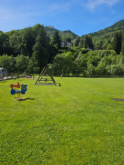 This image depicts a sunny day in the municipality of Pontagna, Italy, specifically in a recreational area with a large, well-maintained grassy field. In the foreground, a red and blue spring rider shaped like a cartoon animal is visible. Beyond that, a wooden A-frame swing set with a black swing is set up on a rubber mat. To the left, a camper van is parked near a wooden fence and benches, suggesting a campground or picnic area. In the middle ground, several people are gathered on picnic blankets in the shade of trees. The background is dominated by a steep, tree-covered mountainside, with houses built into the slopes, indicating a mountainous terrain characteristic of Pontagna. The sky is clear and blue, with a few wispy clouds.