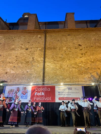This media file captures a lively folk dance performance at the Calella Folk Festival in Calella, Spain. The image is oriented vertically. On stage, a group of performers, dressed in traditional folk costumes, are actively engaged in dancing. The male performers wear white shirts with intricate embroidery, dark pants, and sashes, while the female performers are adorned in colorful dresses with layered skirts and distinct headwear.
The backdrop for the performance is a large banner that prominently displays "Calella Folk Festival." The banner also features the logos of "Ajuntament de Calella" and "FORTUNI VIATGES," along with the website "www.folklorefestival.com." This text clearly indicates the name of the festival, the organizing bodies, and a web address for further information.
The scene appears to be set in the evening or twilight, with the blue sky visible above the stone building to the left. Lighting fixtures are strategically placed, illuminating the stage and the performers. To the right, a spectator is partially visible, holding a smartphone to capture the event, suggesting an audience is present and enjoying the performance. The overall mood is festive and celebratory, showcasing cultural heritage through dance.