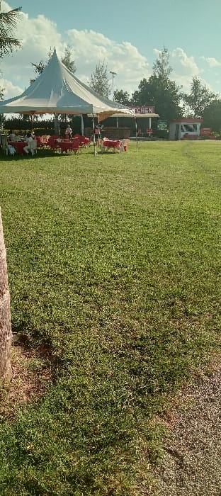 The image shows a grassy field in front of a white tent with red chairs and tables, with a few people standing around. There is a smaller, red building with the word "Kitchen" written on it in the background, along with a few trees. The scene appears to be an outdoor event or gathering. It's located in Athi River, Kenya, specifically at latitude: -1.519, longitude: 36.946.