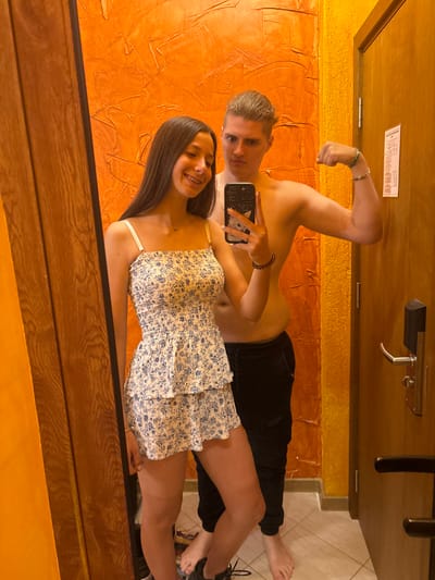 This image is a mirror selfie taken in a room. A young woman with long dark hair and braces is wearing a floral dress and standing next to a young man who is shirtless and wearing black pants. The man is flexing his bicep. The background shows an orange textured wall and a wooden door with a hotel key card lock. The floor is tiled. The location is specified as Kostenets, Bulgaria.
