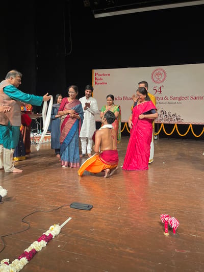 The image shows a group of people on a stage in Chandigarh, India, at the location specified by the provided coordinates. The stage is made of wood and has a wooden floor.  A man is kneeling at center stage wearing a yellow and orange dhoti. He is bare-chested. A man in a white shirt and blue pants is standing behind him. A woman in a red and blue saree stands to the left. She is holding a white scarf. A man in a blue shirt and grey pants is to the right of the kneeling man. Two women in traditional Indian clothing are standing to the right of the man kneeling on the stage. One woman in a red saree is holding a white scarf. The women are part of a group gathered at the stage. Behind the group, there is a large banner with the words “Pracheen Kala Kendra” and "54th Sangeet Sammelan of Indian Classical Arts". The stage is decorated with garlands of white flowers. The background is a dark curtain.