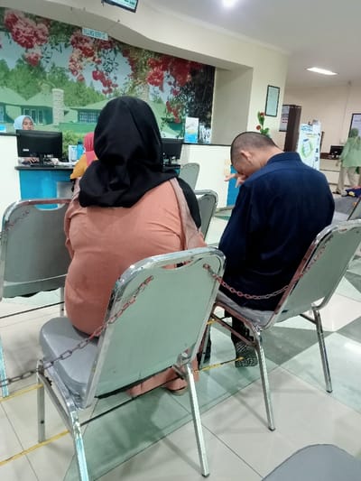 The image shows a waiting area in a building in Kecamatan Margorejo, Indonesia (latitude: -6.76, longitude: 111.015). Two people are sitting on chairs, which are separated by yellow tape and secured together with a metal chain. The woman to the left is wearing a black hijab and peach-colored blouse. She is sitting in a grey chair, with the chair's metal legs and frame visible. The man on the right is wearing a dark blue collared shirt. He is slumped in a chair with his head bent forward, seemingly in thought. He is wearing a black belt and blue jeans. The chairs are in a brightly lit, open space with beige-colored tile flooring. Behind the people is a counter, a partially visible computer screen, and a wall with floral wallpaper depicting a building with an arched entryway. There is a sign behind the counter that reads “Billing Service.” Another sign is visible on the wall to the right of the image. The image is taken from a slightly elevated angle, looking down upon the two people sitting on the chairs.