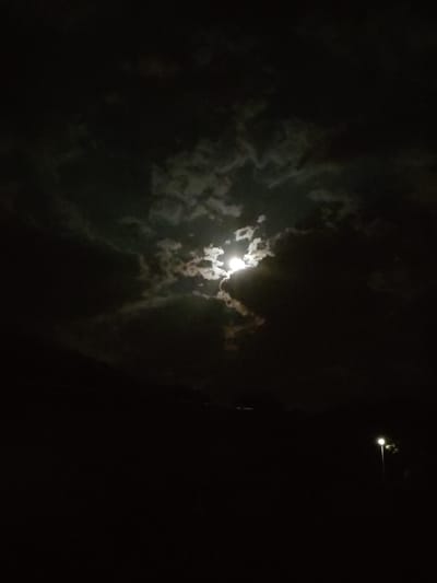 The image depicts a nighttime scene in Tervel, Bulgaria, characterized by a dark sky filled with dramatic, illuminated clouds and the bright moon peeking through. In the foreground, the landscape is predominantly dark, with faint outlines of trees or foliage visible against the night sky. A single street lamp is illuminated on the right side of the frame, casting a soft light onto its surroundings and serving as a point of contrast to the natural illumination of the moon. The overall atmosphere is one of nocturnal beauty and stillness, with no human figures or explicit actions captured. The focus is on the celestial display and the subtle human-made elements present in the urban environment.