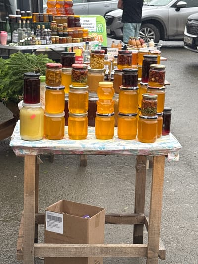 In Inzer, Russia, a market stall displays a variety of jars of honey and preserved goods. The items are arranged on a rustic wooden table covered with a patterned tablecloth. The jars contain honey of different shades of yellow and amber, as well as some with preserved fruits or nuts. Some jars are stacked on top of each other, creating vertical arrangements. In the background, a person is partially visible standing near a vehicle, and other market elements, such as a green banner with text and more bottles on a higher shelf, are present. The ground appears wet, suggesting recent rain. A cardboard box sits beneath the table.