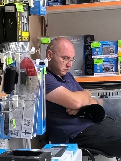 A man with a shaved head and glasses is sitting with his arms crossed, leaning forward, with his head resting on his arms. He appears to be sleeping or resting in an electronics store. The background shows shelves stocked with various electronic items and packaging, including HDMI cables, audio equipment, and other consumer electronics. The scene is well-lit and the items on the shelves are neatly arranged.