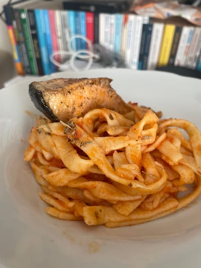 A close-up, eye-level, slightly angled shot captures a white plate filled with pasta and a piece of salmon. The pasta, a wide, flat noodle shape, is coated in a reddish sauce and sprinkled with herbs. A piece of salmon, seared with a dark, crispy skin and a flaky, orange interior, is positioned on top of the pasta. The background is out of focus, showing a bookshelf filled with colorful books. The scene suggests a meal, possibly in a home or restaurant setting. There is no identifiable location in the city of Başakşehir, Turkey, depicted in the image.