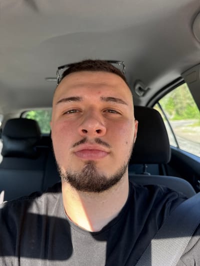 The image, rotated counter-clockwise, features a young man in what appears to be the interior of a car. The man, likely in his 20s or 30s, has short dark hair, a neatly trimmed beard and mustache, dark eyebrows, and brown eyes. He wears a black t-shirt and has eyeglasses resting on his head or collar, partially visible above his left ear. He looks directly at the camera with a neutral to slightly contemplative expression, showing a faint blush on his cheeks.

He is positioned in the driver's or passenger's seat, with a grey car headliner visible above him and a black headrest behind him. A grey seatbelt with a textured pattern crosses the frame, showing sunlight casting a strong shadow on it and his t-shirt, indicating bright, sunny daytime conditions. Through the car's side window, a vibrant green landscape, possibly trees or bushes, is visible, suggesting an outdoor, natural environment. The scene takes place during the day, under clear skies. There is no visible text in the image. Given the provided context, the setting is Laka, Bulgaria.