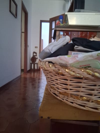 The image shows an interior scene, likely a home in Colorno, Italy. A woven basket filled with various items, including what appears to be clothing and packaging, is prominently displayed. The basket has a decorative golden ribbon woven through it. To the left of the basket, a wooden cabinet with drawers is visible. A white appliance, possibly a refrigerator or washing machine, is partially visible on the far left. In the background, a hallway leads to a door, and there's a glimpse of a tiled floor. A small framed object is on the wall to the left, and a light switch is visible in the distance. The overall impression is of a domestic space with items stored in a basket. There is no discernible activity or event taking place; it appears to be a still life composition. The lighting suggests it is daytime, but no specific time of day or weather conditions can be determined. Text is visible on one of the items in the basket, appearing as "CONAD" printed on a green and white package.