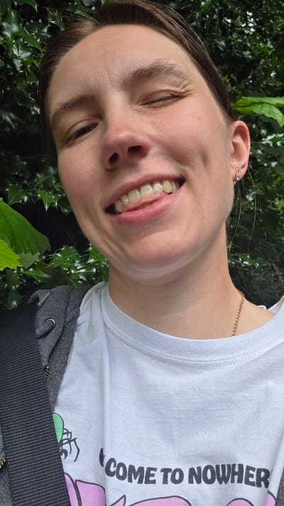 A close-up, selfie-style portrait of a young person with light skin and brown hair is shown, positioned outdoors in a garden setting. The individual is winking, with their tongue slightly protruding from their mouth. They are wearing a white t-shirt with text and graphics, and a grey hooded jacket. A dark strap crosses their chest. The background consists of lush green foliage. The image is taken in Portland, Oregon.