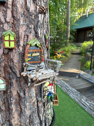 This image depicts a detailed miniature fairy village attached to the trunk of a large tree. The village includes tiny houses, windows with green leaf-shaped frames, a "Welcome" sign, and a small wooden balcony. A gnome figure is visible inside a tiny enclosure, and a decorative bee figurine is positioned as if flying towards the village. The tree is set in a natural environment with a wooden pathway and a rustic log cabin visible in the background, suggesting a location in a rural or forested area. The presence of a green, artificial turf-like ground cover indicates a manicured garden setting. The overall scene is whimsical and intricately designed, creating a charming miniature world. The location in Sukhaya, Russia, implies a specific geographical context for this garden installation.
