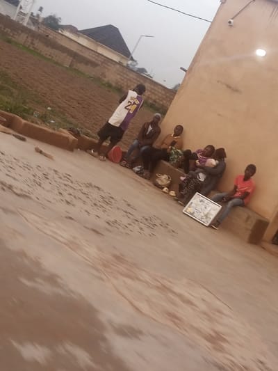 This media file shows a group of people gathered outdoors, possibly in Kwang, Nigeria. The scene appears to be a residential area with a building made of light-colored material, a dirt or field area, and a brick wall. The time of day seems to be overcast, possibly morning or late afternoon, due to the diffused light and muted colors.

The subjects include several individuals, some sitting and others standing. One person stands out wearing a purple and white basketball jersey with the number 24 on the back. Several people are seated on a raised concrete structure or bench in front of the building. Two individuals in the center appear to be holding and interacting with a baby. Another person is seated further to the right, wearing a red t-shirt. A framed picture or poster is propped up on the ground in front of the seated group. A red bucket and some bags are also visible on the ground.

The overall mood seems relaxed, with people gathered together, though it's difficult to discern specific emotions due to the image quality and distance. The setting suggests a casual outdoor gathering, perhaps a family or community get-together. The building has a security camera and a light fixture mounted on its exterior. The ground is a mix of paved and unpaved areas, with some visible puddles or damp patches. The distant background shows a dark-roofed structure and some vegetation.