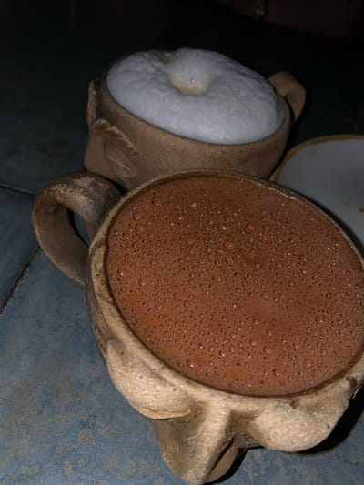 Two mugs are shown on a blue tile surface, likely in Oaxaca City, Mexico. The mug on the left contains a white frothy liquid, possibly atole blanco. The mug on the right contains a brown, bubbly liquid, likely chocolate or a similar drink. Both mugs appear to be made of a textured, light-colored material, possibly clay. The background is dark and out of focus. A small portion of a white plate is visible. The lighting suggests the image was taken indoors with artificial light.