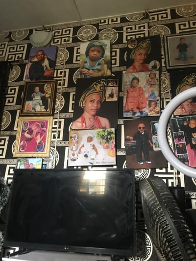 The image displays an interior wall within a residence in Oba-Ile, Nigeria, densely covered with various framed and unframed photographic prints and canvases. The wall is adorned with black and white wallpaper featuring a repeating geometric pattern composed of interlocking lines and circular medallions.
Numerous portraits of individuals are mounted on the wall. These include multiple adult females, often depicted wearing traditional Nigerian headgear (gele) and beaded necklaces, some of these prints incorporating smaller inset photographs of children or alternative poses. Portraits of adult males and numerous children and babies are also visible. Specific examples include a baby in a white outfit with "Abdul Hafeez @ 3months" inscribed below, a child in traditional attire, and a child wearing a suit and sunglasses. Several photographs are enclosed in ornate gold-colored frames, while others are canvas prints. A light-colored, semi-circular decorative object is affixed to the upper left portion of the wall, and a horizontal wire with small clips is suspended across the upper wall section.
Positioned beneath the wall display is a black LG flat-screen television, whose screen exhibits smudges and reflects an overhead light source or window. To the right of the television, the gridded, dusty back of a black electric fan is partially visible, angled upwards. A green, tinsel-like decorative element is partially observable to the left of the television.