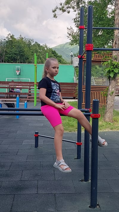 A young girl with blonde hair pulled back in a ponytail is performing an exercise on a set of parallel bars outdoors. She is wearing a black t-shirt with a graphic print and bright pink cycling shorts. Her left leg is extended forward, resting on the upper bar, while her right leg is bent, with her foot on the lower bar. She is wearing white sandals on both feet. Her gaze is directed away from the camera, and her expression is neutral.
The parallel bars are dark gray with red connectors. The ground beneath the bars is covered with dark gray rubber matting. In the background, there is a wooden structure with horizontal planks and a green building, possibly a sports facility, with a basketball hoop visible. Trees and a cloudy sky are also in the background, with rolling hills in the distance, suggesting a rural or mountainous setting. The image appears to be taken during the daytime, under overcast skies. The overall atmosphere is that of a public outdoor fitness area or park.