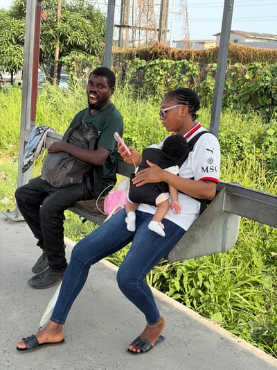 The image depicts an outdoor scene in Jakande, Nigeria, featuring two adults and an infant seated on a metal public bench under bright daylight. The background consists of dense green vegetation, with utility poles and distant buildings partially visible above the foliage. The ground beneath the bench is concrete.
On the left, a dark-skinned male with a beard and short dark hair is seated, leaning back against the bench. He wears a dark green t-shirt, dark trousers, and dark closed-toe shoes. A dark backpack rests on his lap, draped with a light-colored fabric. His mouth is wide open, revealing his teeth, and his gaze is directed towards the right side of the frame.
To his right, a dark-skinned female with braided hair and pink-rimmed glasses is seated, wearing a white short-sleeved t-shirt with black and red collar detailing and sponsor logos including "PUMA" and "MSC." She wears blue denim jeans and black open-toed flat sandals, with a gold-colored ankle bracelet on her right ankle. She holds a light pink smartphone in her left hand, which appears to be connected to a white charging cable, and she is looking down at the device. An infant with dark curly hair, wearing a white top and white booties, is secured in a black baby carrier on her chest, facing away from the viewer. A pink bag is positioned on the bench between the two adults, and a white umbrella is leaning against the female's legs. No direct interaction between the adults is observed; each individual is engaged in their own activity or expression.