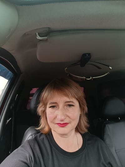 A medium shot captures a middle-aged Caucasian woman with auburn hair and light skin, wearing bright red lipstick and a black, ribbed shirt, seated in the driver's seat of a car. She has freckles scattered across her nose and cheeks and wears eyeliner and mascara. Her left eye is open and looking at the camera, while her right eye is partially visible. A pair of sunglasses hangs from a clip on the car's ceiling, just above her head. The interior of the car is visible, with black leather seats and the car's headliner. The lighting suggests it is daytime, with natural light illuminating the scene from the front and left. The image appears to have been taken inside a vehicle.