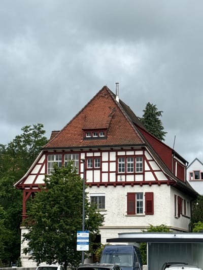 An image taken in Waldshut, Germany, captures a traditional German house with a half-timbered facade under an overcast sky. The house, situated on the left side of the frame, features white stucco walls with prominent dark red wooden beams forming a half-timbered pattern. It has a steeply pitched roof covered in reddish-brown clay tiles, with several small dormer windows protruding from the roofline. A metallic chimney is visible near the peak of the roof.
On the upper floor of the house, there are windows with dark red shutters, some open and some closed. The lower part of the house is partially obscured by dense green foliage from trees and bushes. To the right of the house, parts of two vehicles are visible: a dark blue car (possibly a van or SUV) and a lighter-colored vehicle above it.
A blue and white street sign is prominently displayed, indicating parking regulations. The sign clearly shows a white "P" symbol and the text "1 Stunde" (1 hour), suggesting a one-hour parking limit. Further text on the right side of the sign is less distinct but appears to outline additional parking rules, possibly mentioning a "Parkscheibe" (parking disc).
The scene is set during daytime, characterized by the muted light filtering through heavy gray clouds, indicative of an overcast or potentially rainy day. There are no people visible, and no specific activity is taking place. The architecture and the street sign clearly identify the European, specifically German, context of the image.