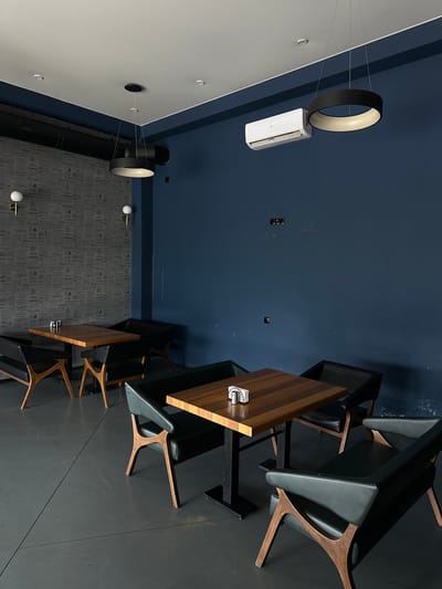 A bird's-eye view shows the interior of a contemporary establishment in Marneuli, Georgia. The ceiling is partitioned into a white section, a deep blue segment, and a patterned gray portion. A ring-shaped lamp hangs suspended near an air conditioning unit. Below, wooden tables are arranged alongside gray chairs with wooden frames. The floor alternates between patterned gray and a solid dark shade. A dark, vertical pipe runs along the wall from floor to ceiling. The scene is devoid of people.
