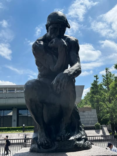 A bronze sculpture of Auguste Rodin's "The Thinker" is centrally positioned on a stone plinth, viewed from a low angle against a bright blue sky with scattered white clouds. The muscular, nude male figure is seated, with his right elbow resting on his left thigh, and his chin supported by his right hand, gazing downwards. The sculpture exhibits a dark, patinated surface.

In the background, behind and to the left of the statue, a modernist concrete or stone building with large, rectangular dark-paned windows is visible. This structure is identified as part of the National Museum of Western Art, located in Ueno Park, Taito, Tokyo, Japan. A strip of green foliage or hedges runs along the base of this building. To the right and further back, dense green trees indicate a park environment, with additional geometric concrete or stone structures visible amidst the greenery.

In the mid-ground, a black metal fence with vertical bars and diagonal cross-elements extends horizontally across the lower portion of the image. To the left of the statue, behind this fence, a person in dark clothing, including a backpack, walks to the right. A single, spherical white light fixture on a dark pole stands behind the fence in this area. To the right of the statue, also behind the fence, the upper torso of another person wearing a light-colored shirt and a dark baseball cap is partially visible, looking downwards. The ground directly beneath the statue's plinth appears to be paved with coarse, light-colored stone.
